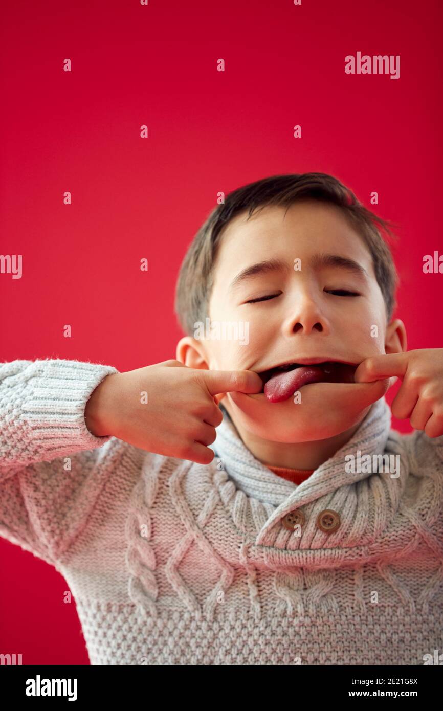 Portrait Of Young Boy Against Red Studio Background Pulling Funny Faces At Camera Stock Photo