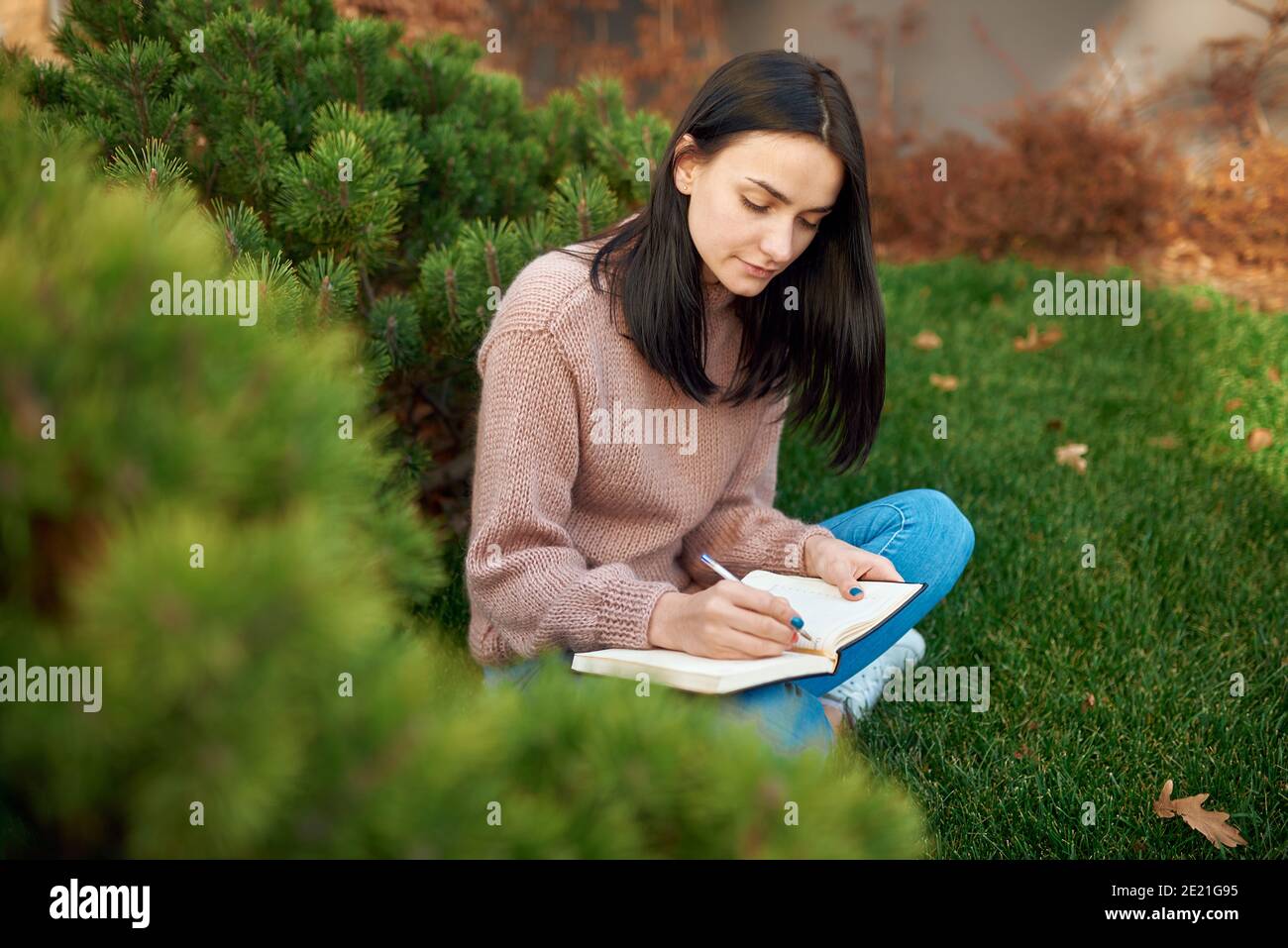 Pleasant young brunette with long hair is concentrated on writing on page of her notebook outside on an open lawn Stock Photo