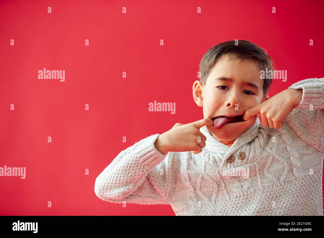 Portrait Of Young Boy Against Red Studio Background Pulling Funny Faces At Camera Stock Photo