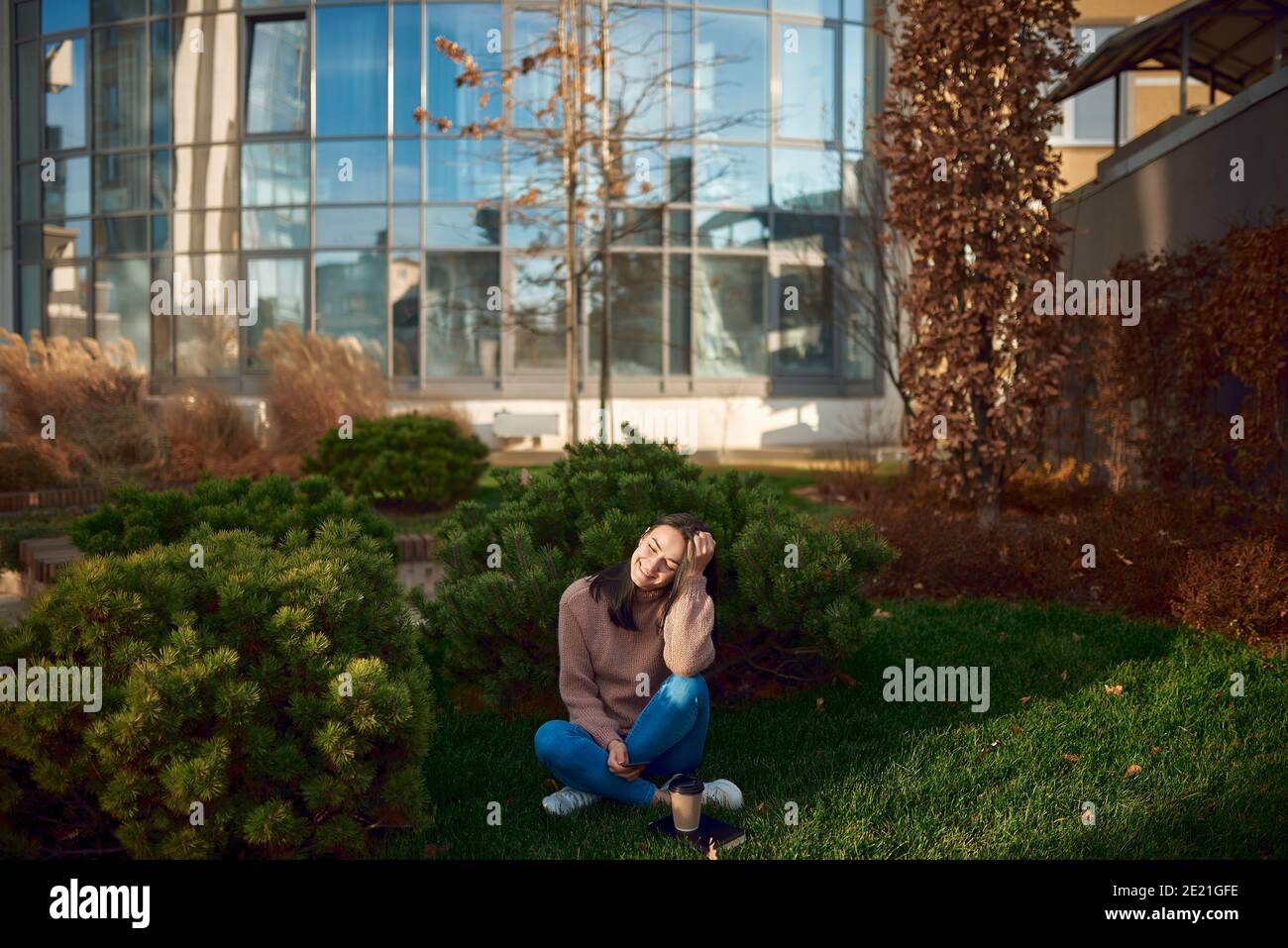 Lovely young lady feeling glad looking on screen of her telephone, posing on grass lawn close to tall modern construction Stock Photo