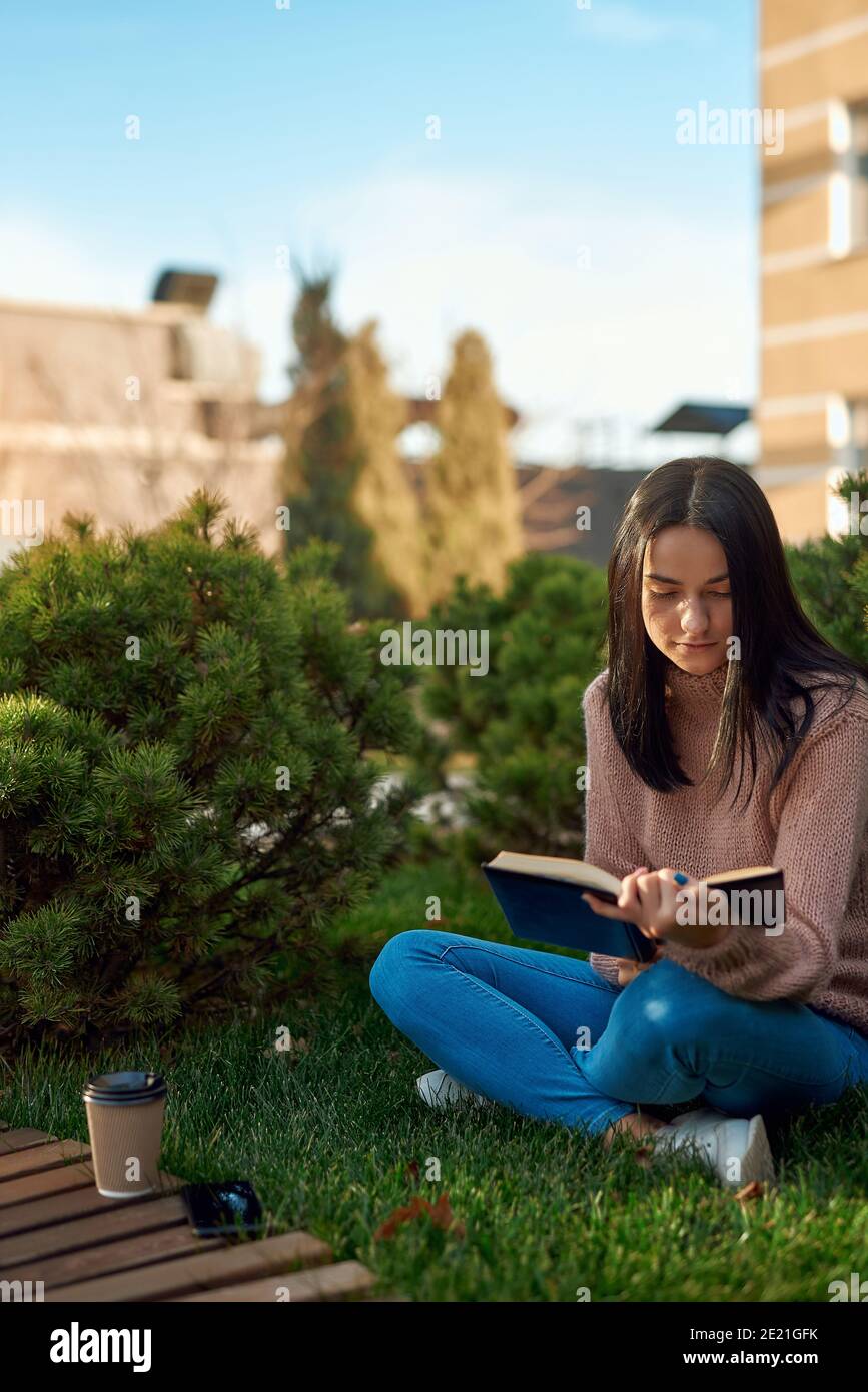 Pensive young lady enjoying interesting book while sitting on grass in a frontage before a tall modern building Stock Photo