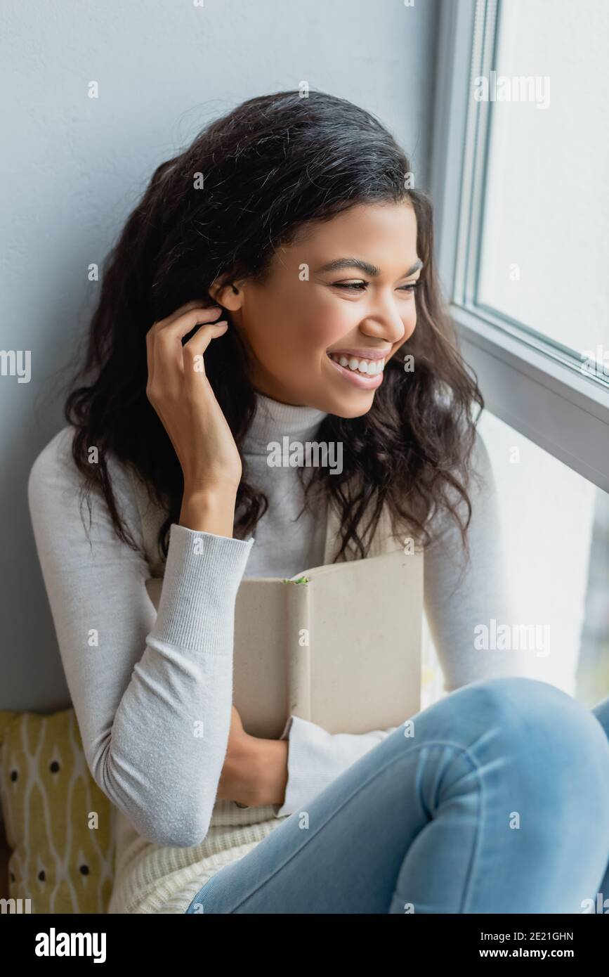 cheerful african american woman fixing hair and looking through window while holding book at home Stock Photo - Alamy cheerful african american woman fixing hair and looking through window while holding book at home Stock Photo - Alamy