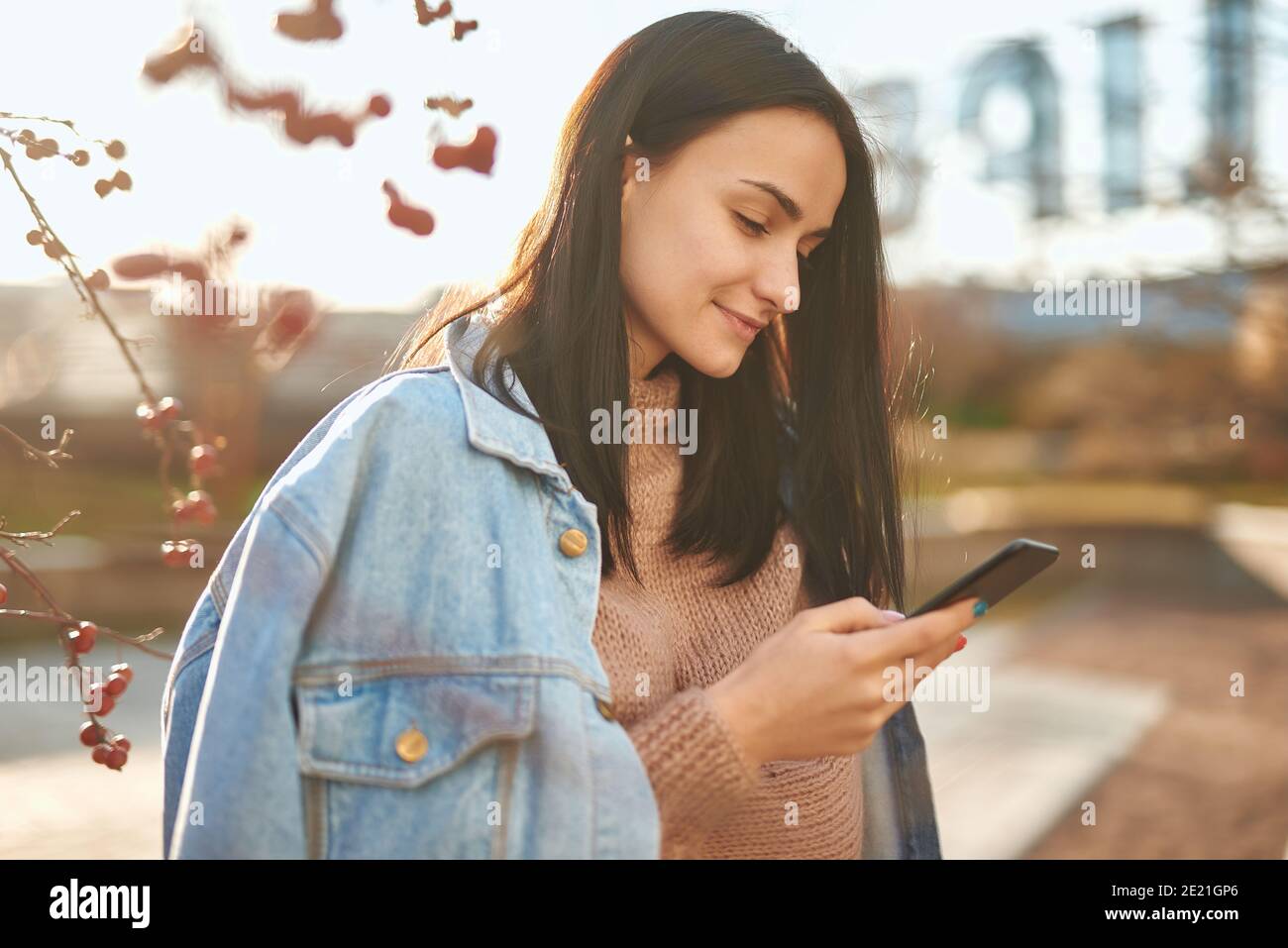 Side shot of a smiling young lady in casual clothing messaging by her cellphone on blurred natural background Stock Photo