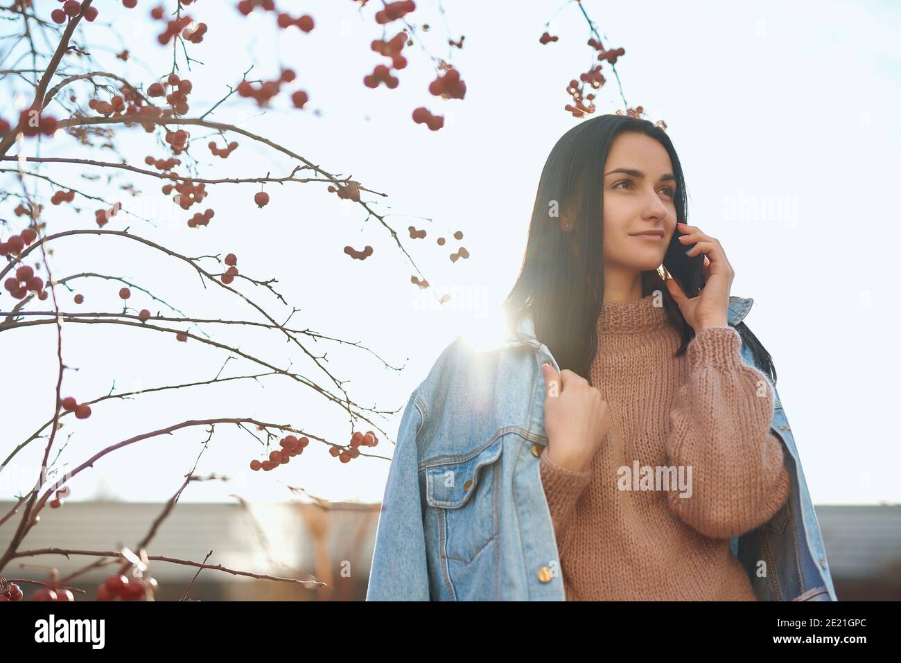 Adorable lady walking outdoors in a jeans jacket over her shoulders while communicating by mobile phone Stock Photo