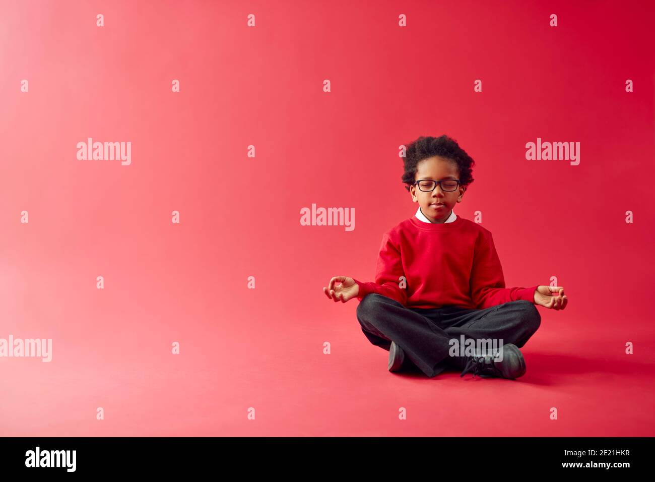 Male Elementary School Pupil Wearing Uniform Sitting And Meditating Against Red Studio Background Stock Photo
