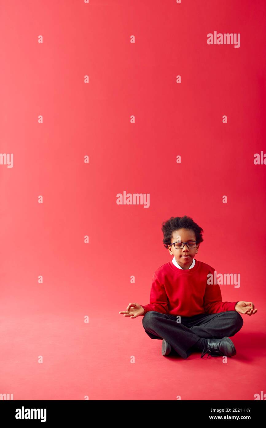 Male Elementary School Pupil Wearing Uniform Sitting And Meditating Against Red Studio Background Stock Photo