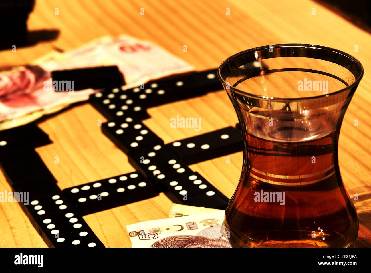 Tea glass with black tea stands next to a domino on a pile of Turkish paper money on a grained wooden board Stock Photo