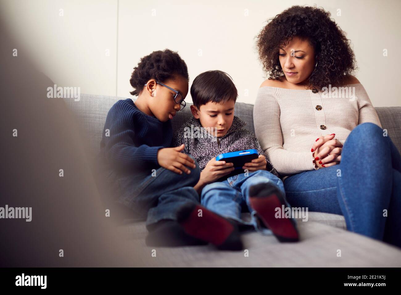 Mother And Sons Sitting On Sofa At Home Playing Computer Game Together On Hand Held Device At Home Stock Photo