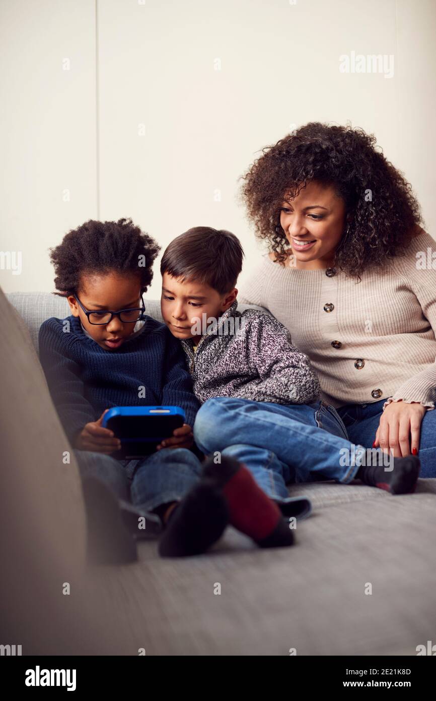 Mother And Sons Sitting On Sofa At Home Playing Computer Game Together On Hand Held Device At Home Stock Photo