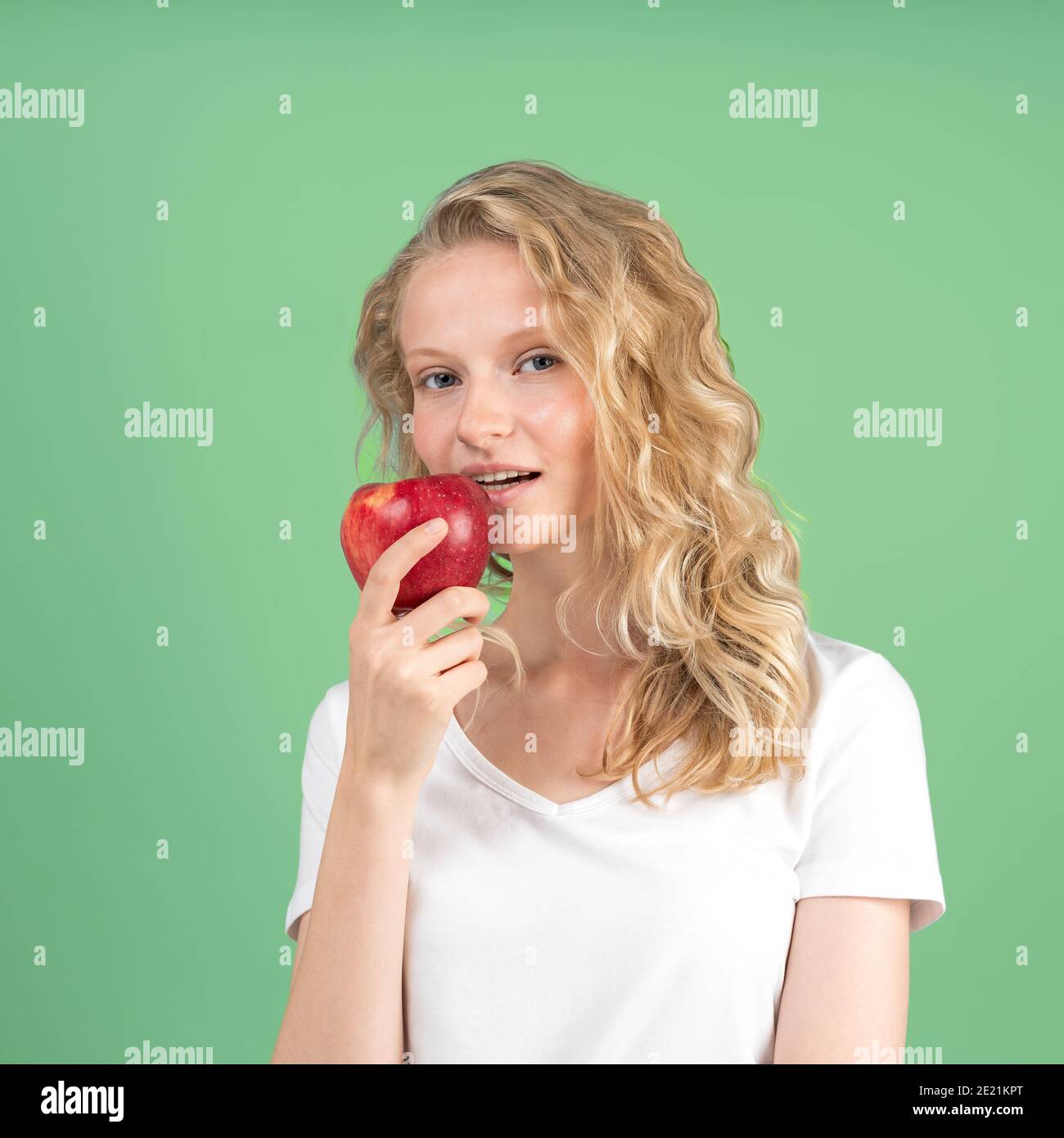 Portrait of young smiling woman bitting red apple againt green wall background. Fresh face Stock Photo
