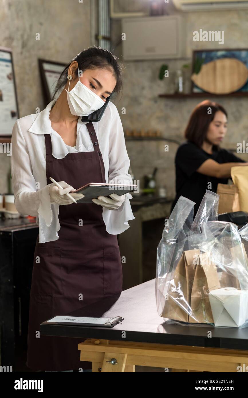 Asian waitress take order from mobile phone for takeout and curbside pickup orders while city lockdown. They wear protective face mask to prevent from Stock Photo