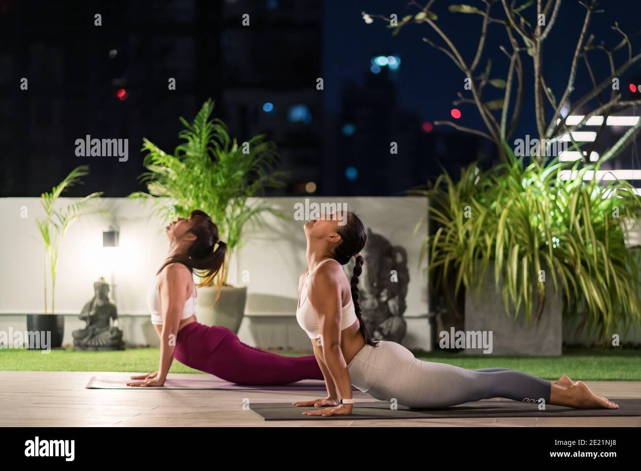 Two asian women in sportswear pants practicing yoga during city lockdown in their apratment with distance in background of cityscape illumination at n Stock Photo