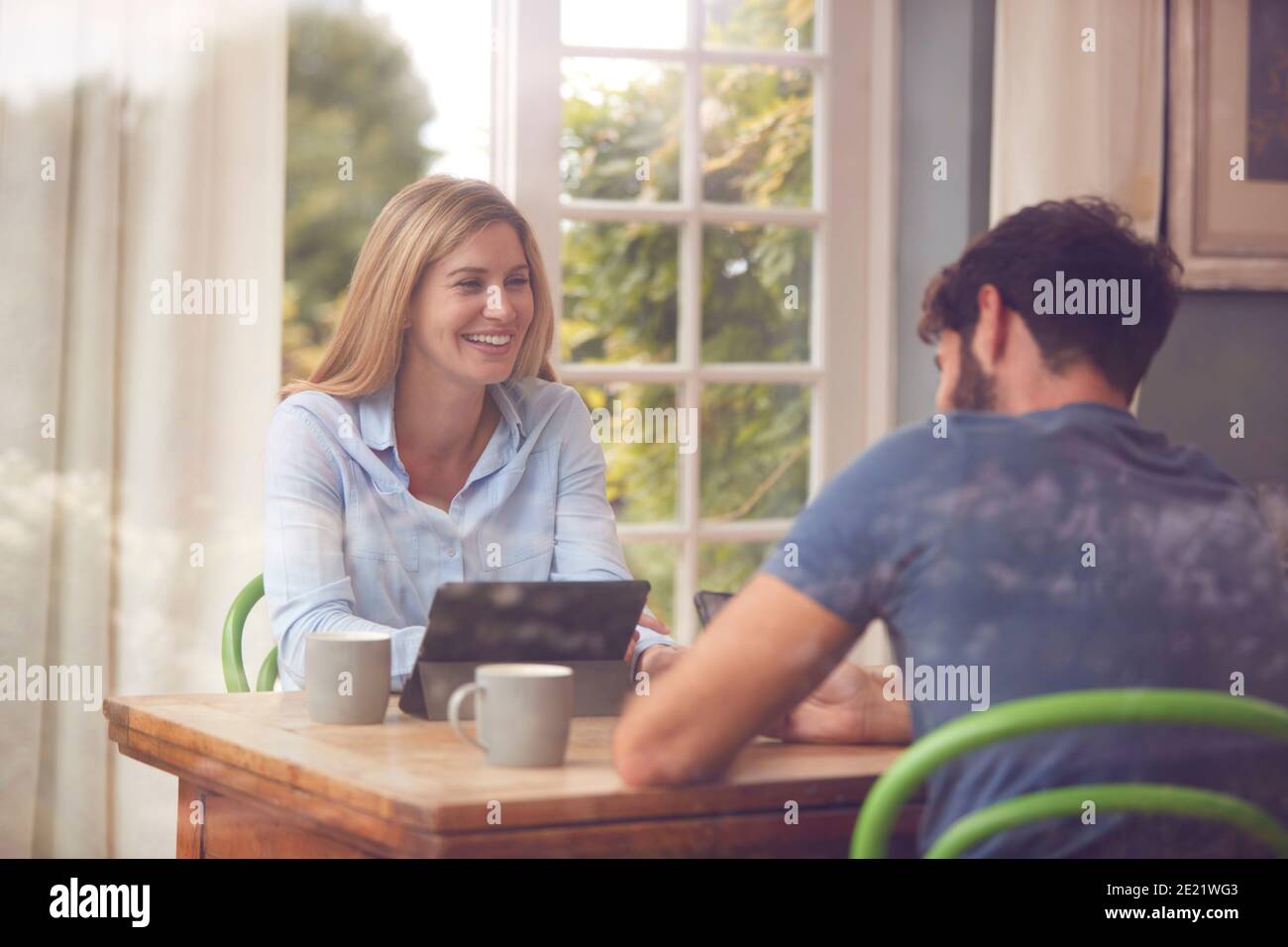 Couple With Digital Tablet Sitting At Table Working From Home Viewd Through Window Stock Photo