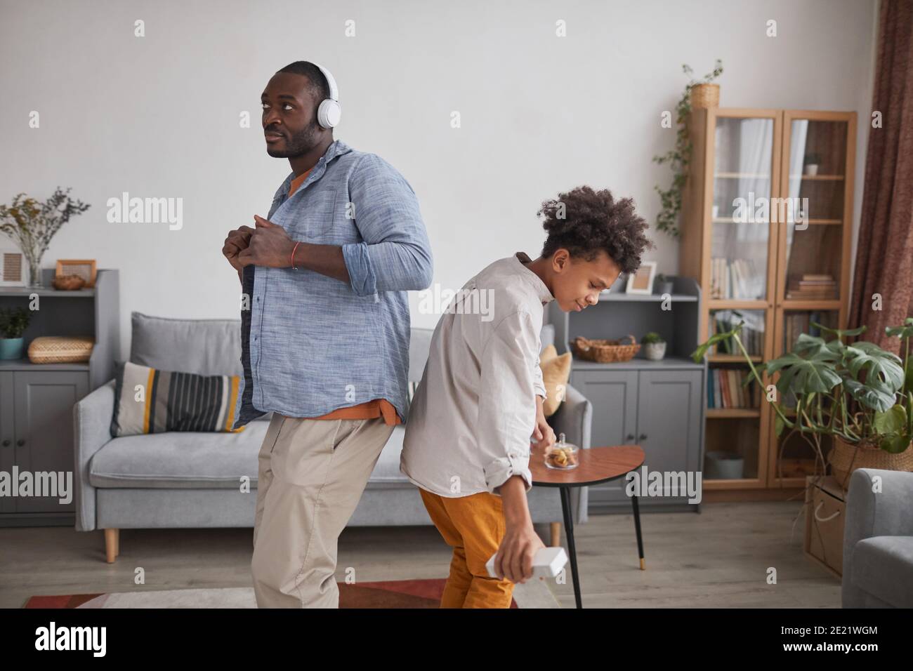 Medium long shot of joyful African American man and teen boy having fun dancing in living room at home Stock Photo