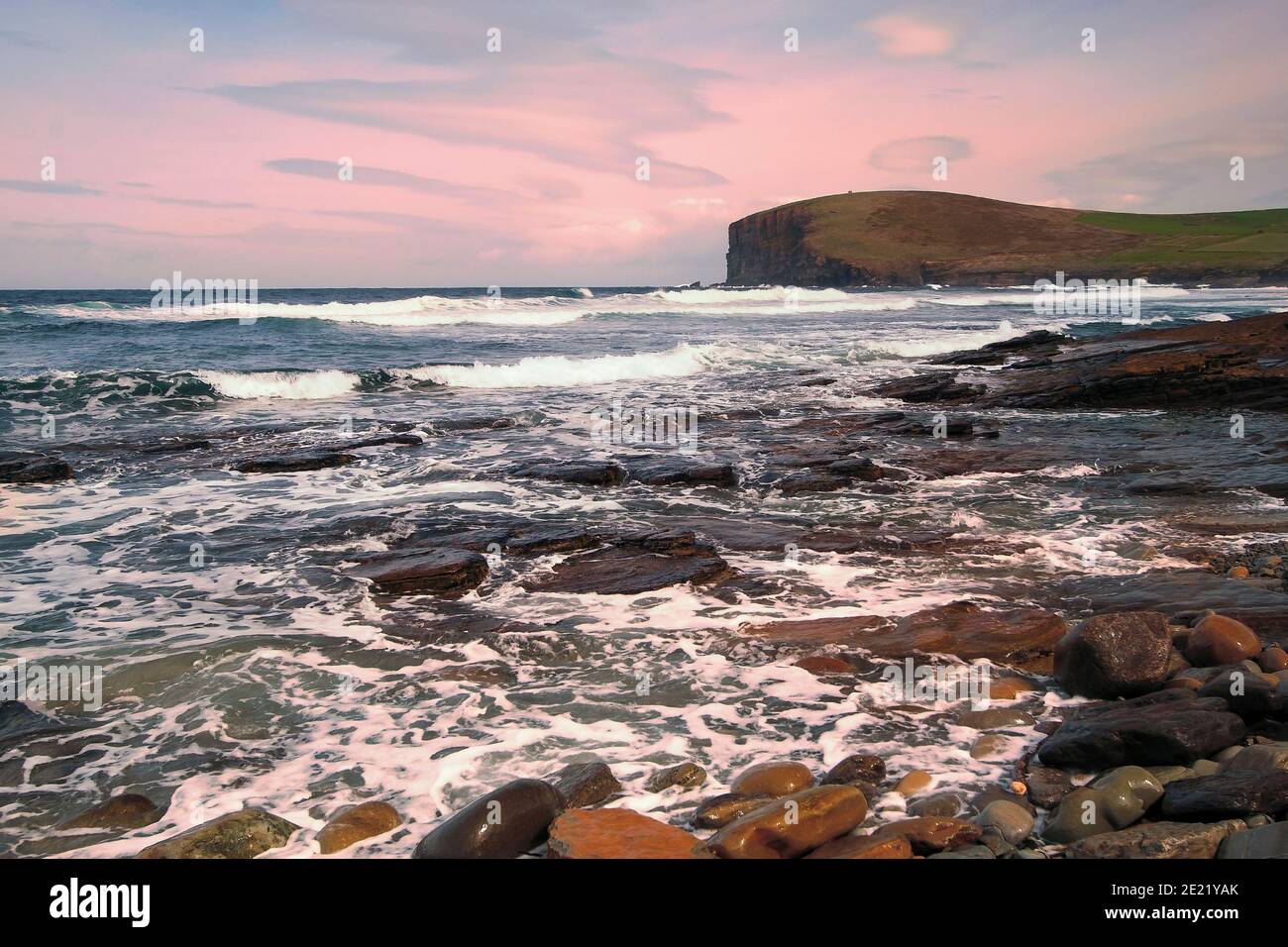 Rough sea waters on west shoreline of Orkney islands with stones on the beach and cliffs in background Stock Photo