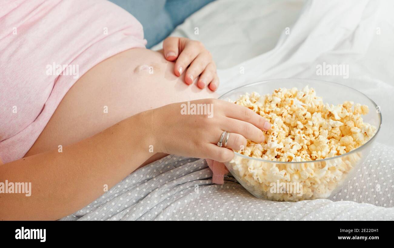 Closeup of pregnant woman in pajamas relaxing in bed and eating popcorn
