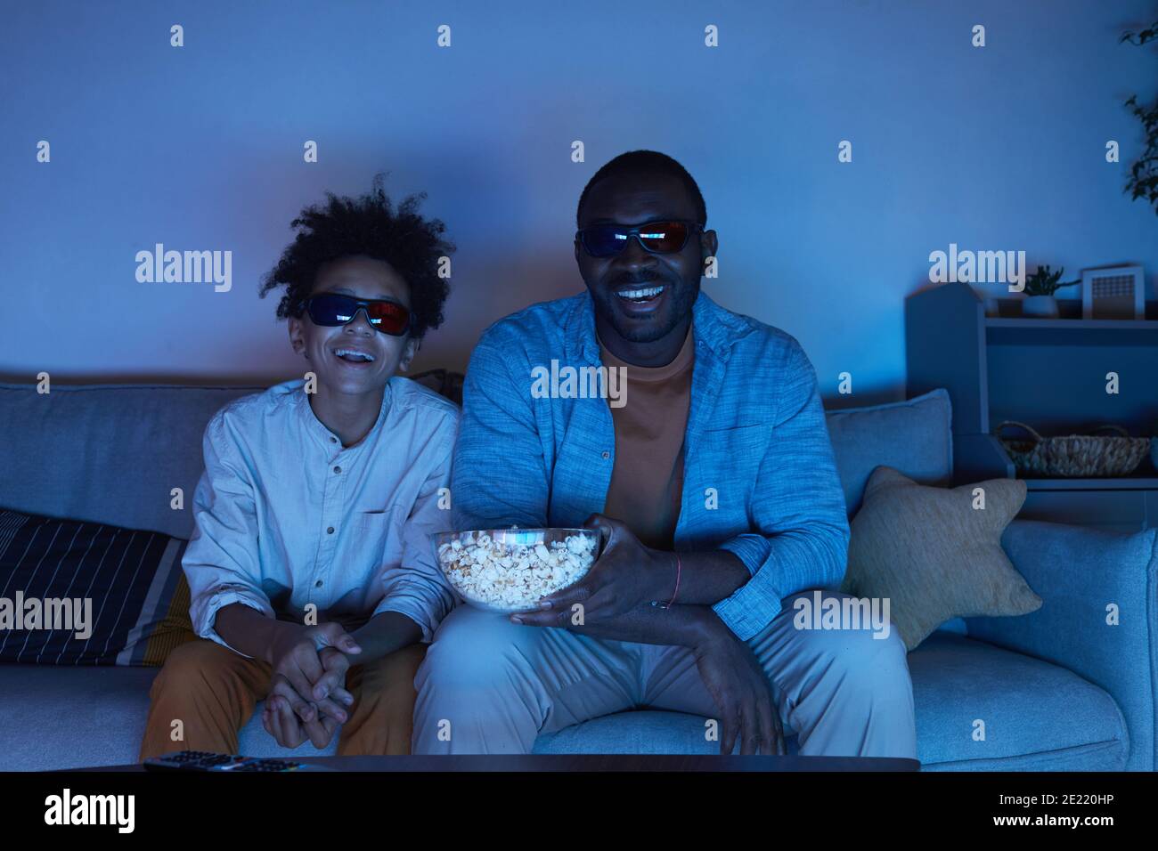 Portrait of African American man and his teen son sitting together on sofa in living room laughing while watching 3D movie and eating popcorn Stock Photo