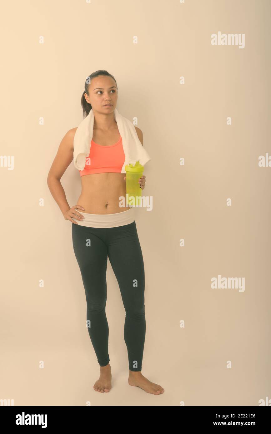 Full body shot of young Asian woman thinking while looking up and holding water bottle with hand towel around neck ready for gym against white Stock Photo