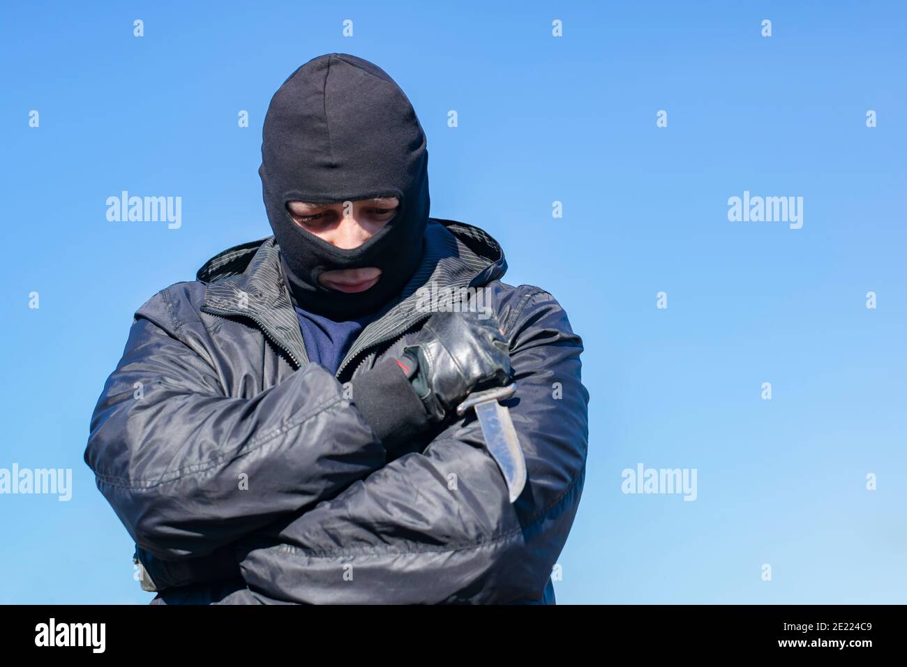 a sad, thoughtful bandit stands in a black mask and with a knife against the blue sky Stock Photo