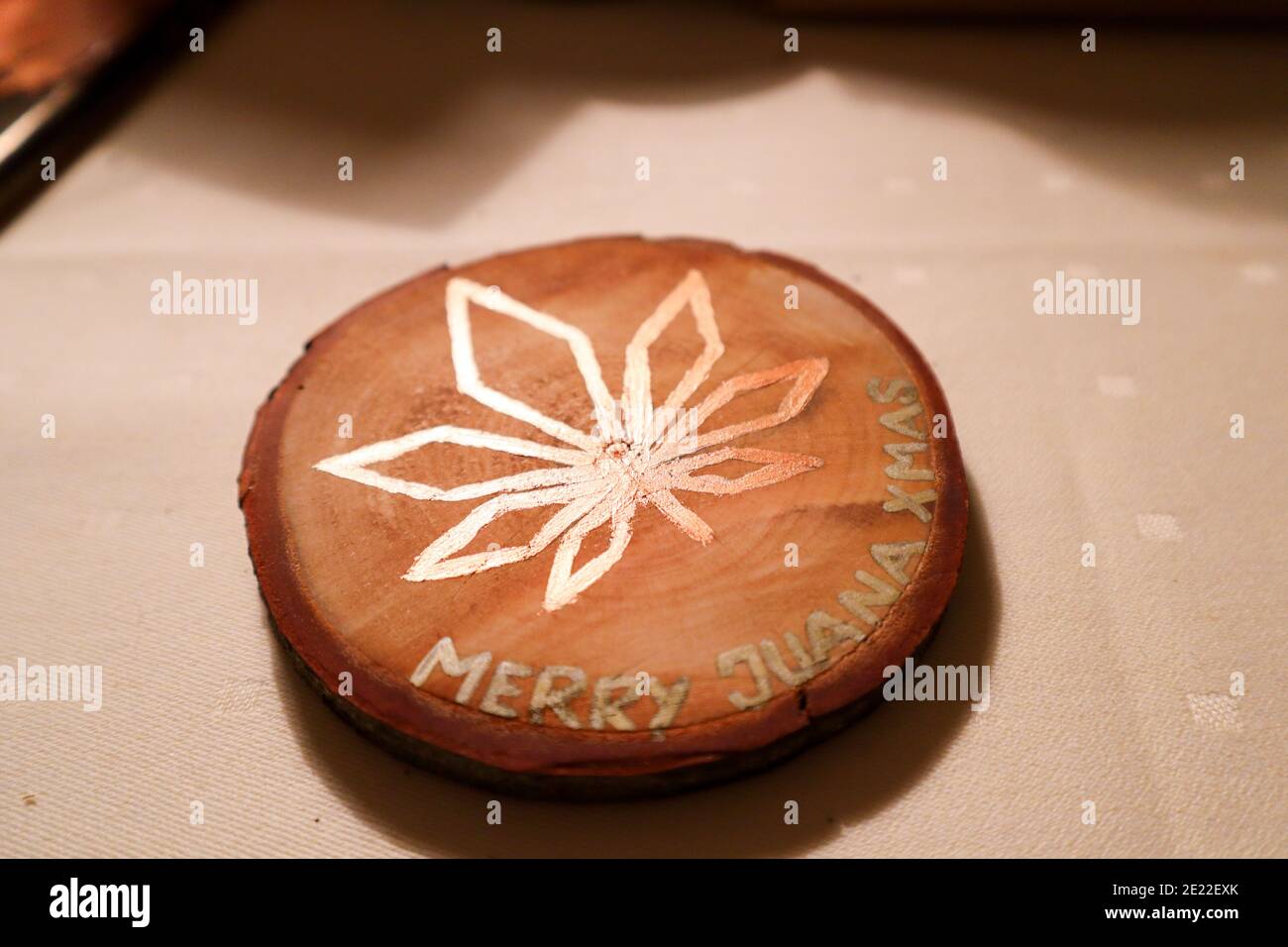 Selfmade wooden coaster on a table with a marijuana leaf in bright colors. The text MERRY JUANA XMAS decorates the coaster. Stock Photo