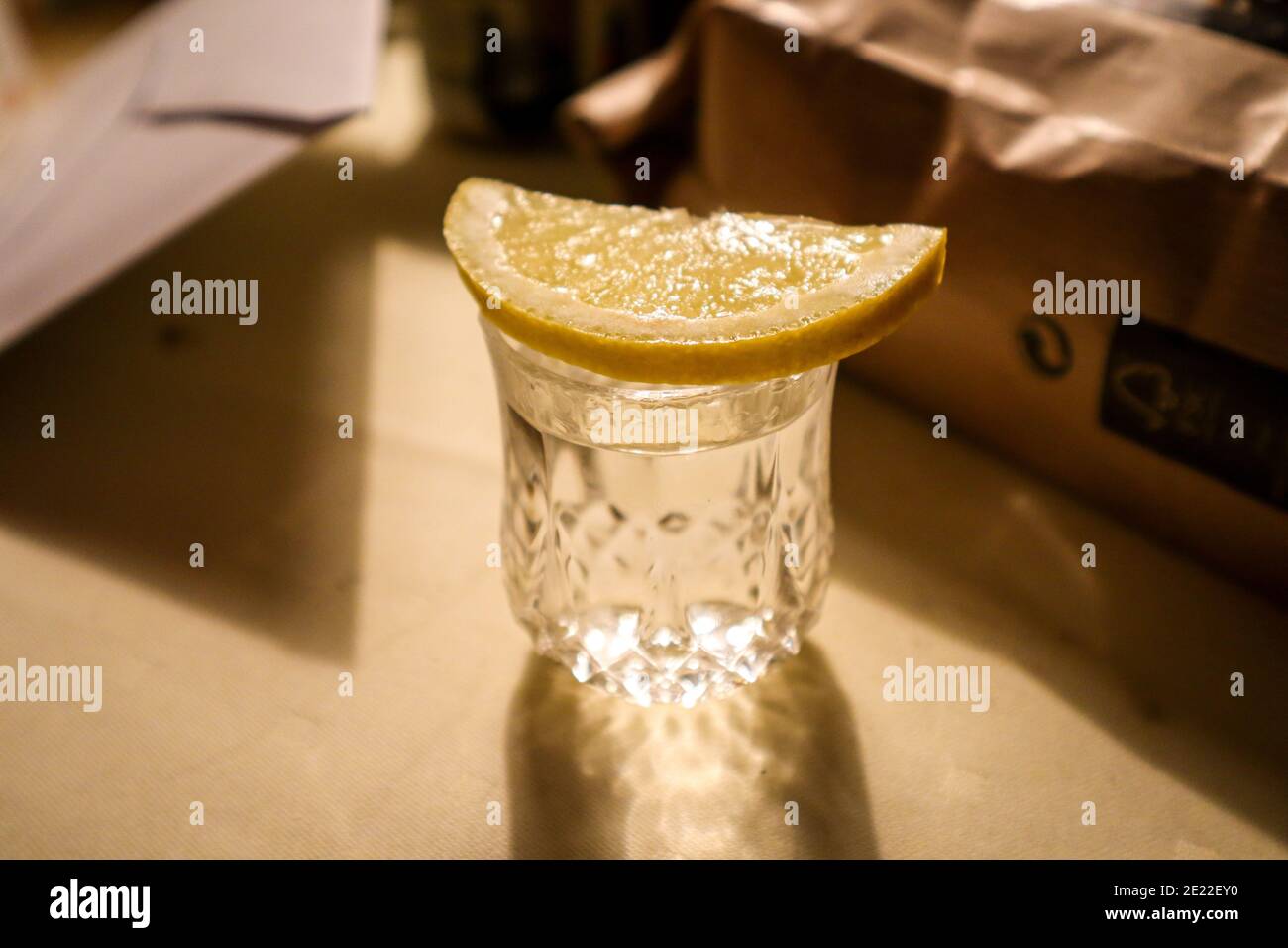 Tequila shot glass with freshly sliced lemon on a table with great lighting which shines through the glass. There are objects in the background. Stock Photo