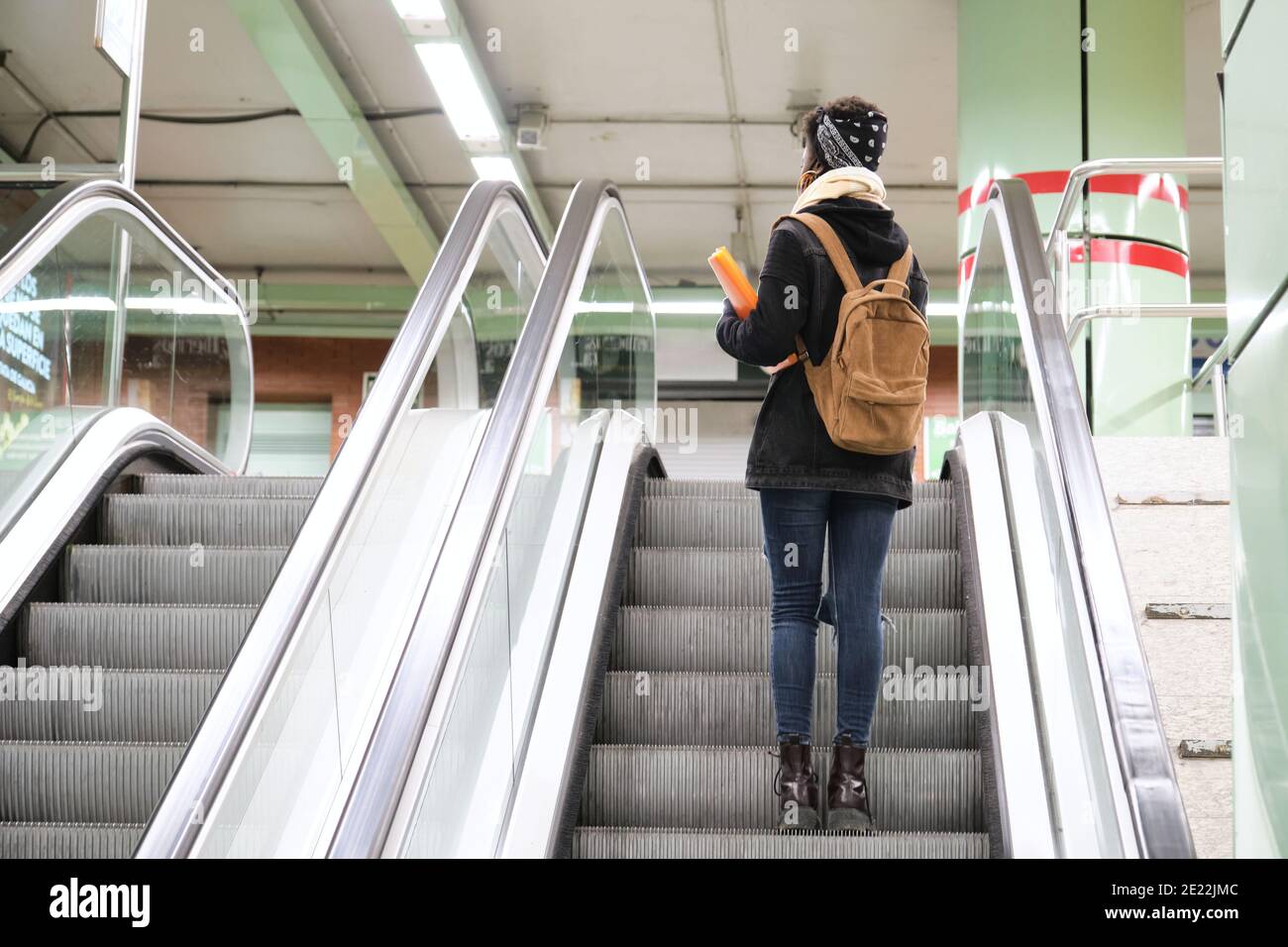 university female african student wearing protective face mask taking escalator upstairs at the underground station. New normal in public transport. Stock Photo