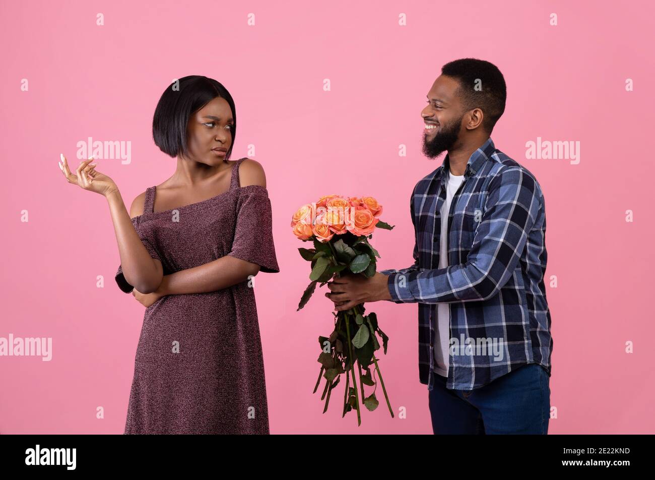 Irritated black woman rejecting geek man offering flowers on Valentine's Day over pink studio background Stock Photo