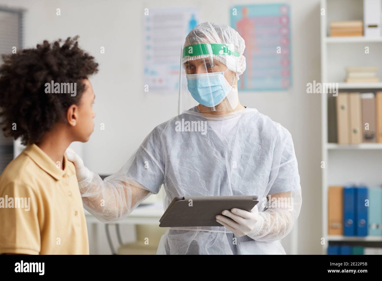Portrait of doctor wearing full protective gear talking to African-American boy during consultation in clinic, copy space Stock Photo