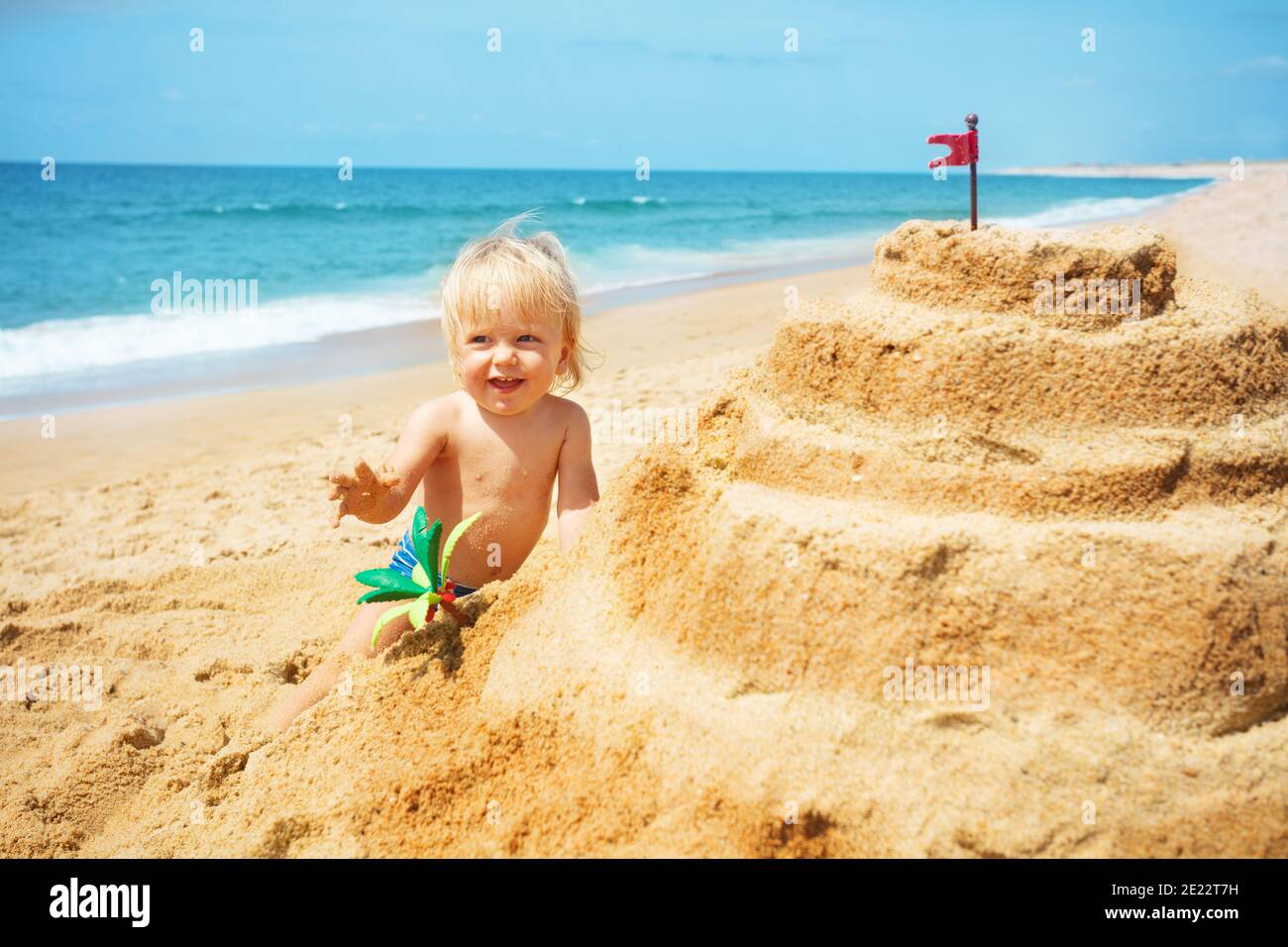 Happy little blond boy building sand castle on beach near the sea wave hand and smile Stock Photo