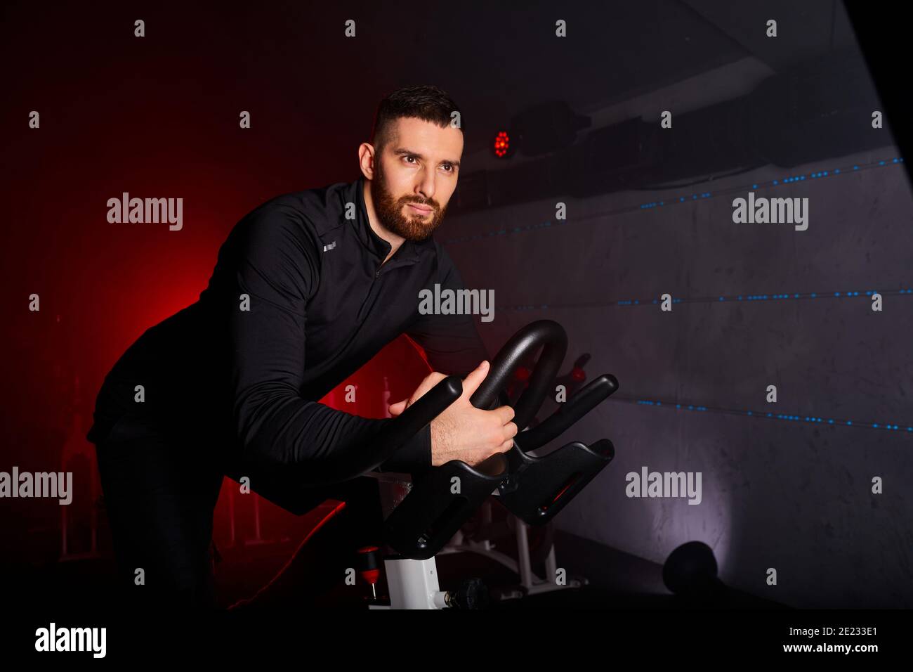 sportsman training at sport equipment in the gym, confident and strong male in tracksuit has intensive training on bicycle in fitness club, looks at c Stock Photo