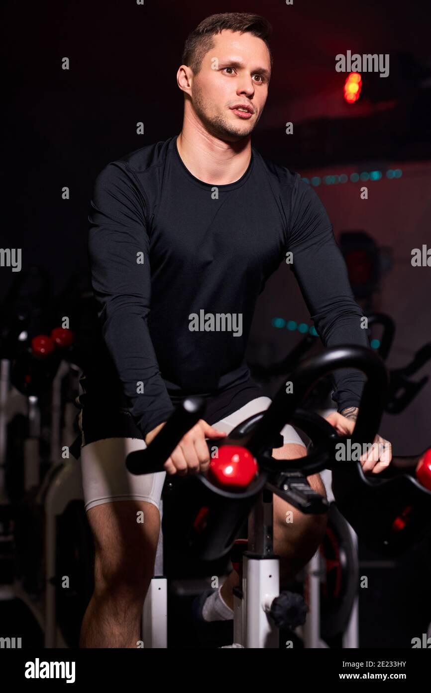 man training at sport equipment in the gym, confident and strong male in tracksuit has intensive training on bicycle in fitness club Stock Photo