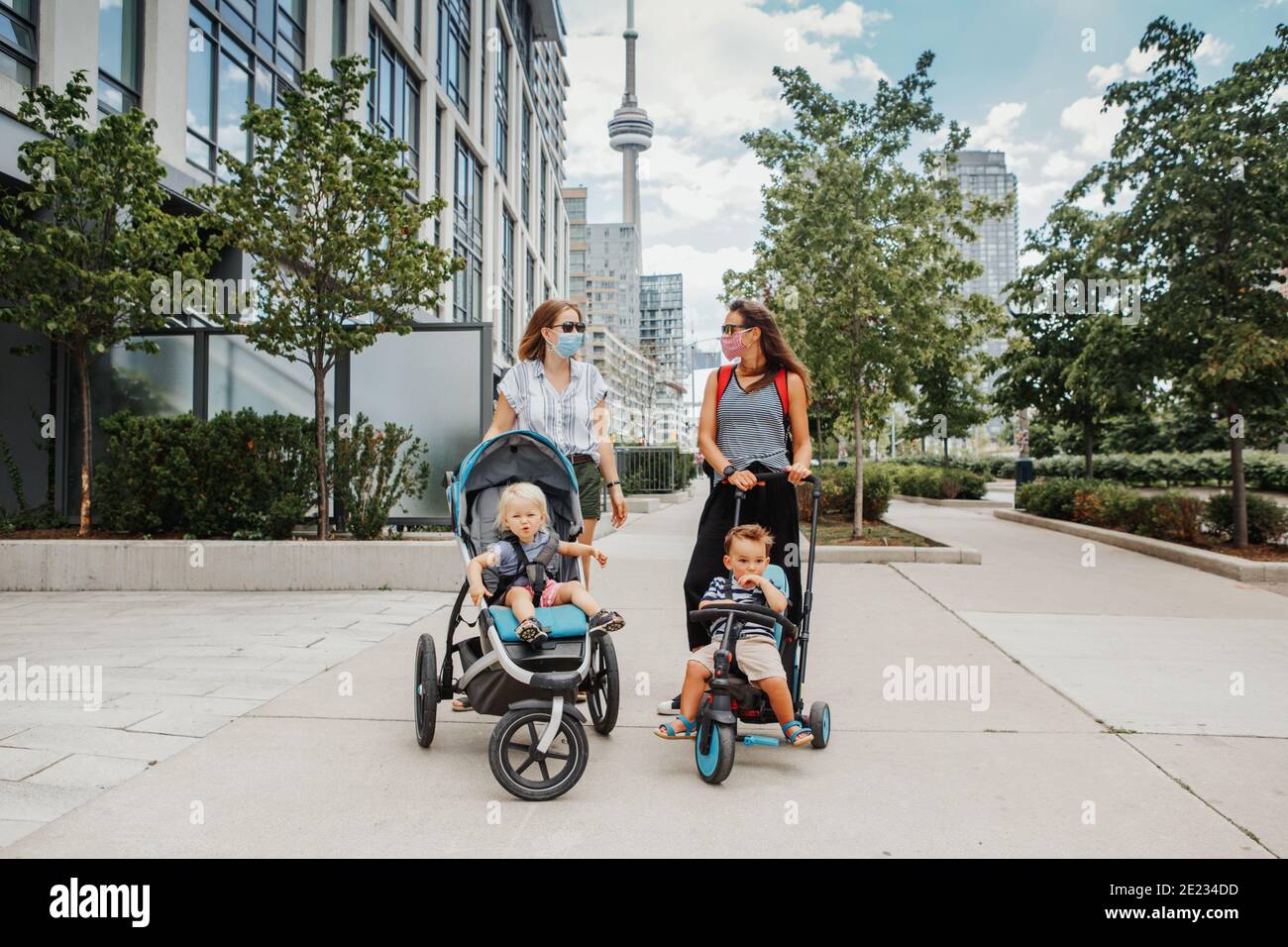 Two Caucasian moms with strollers and kids walking together in Toronto city Canada. Women in face masks with children outdoor. Friends talking Stock Photo