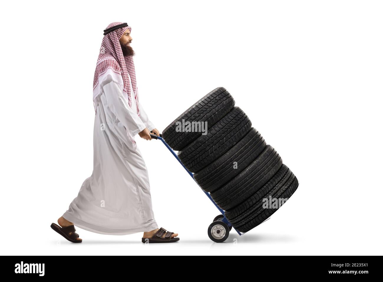 Full length profile shot of a saudi arab man wearing a thobe and pushing a hand truck with a tires isolated on white background Stock Photo