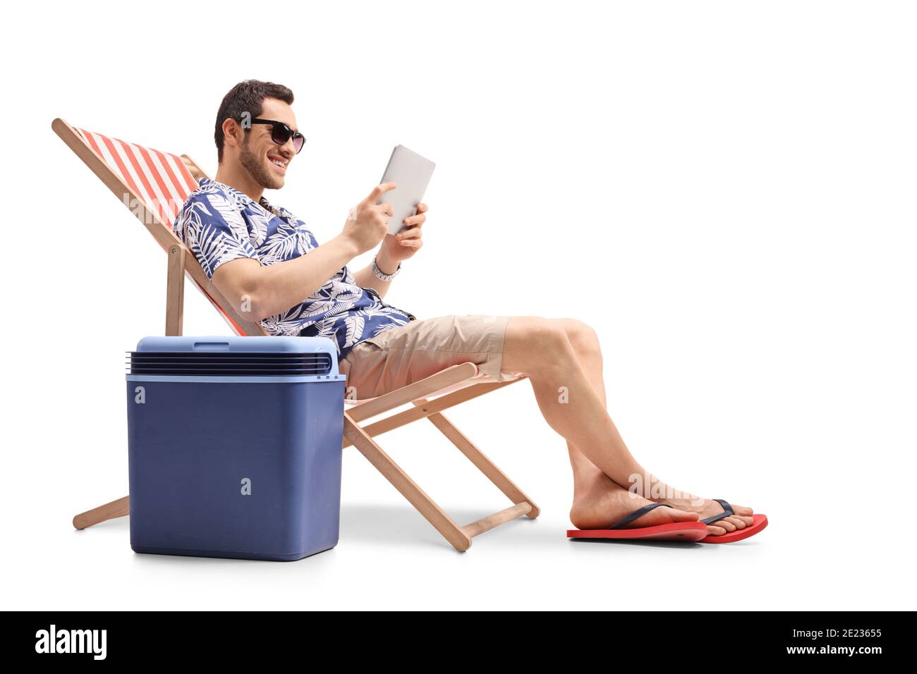 Young man sitting in a deck chair next to a cooling box and holding a digital tablet isolated on white background Stock Photo