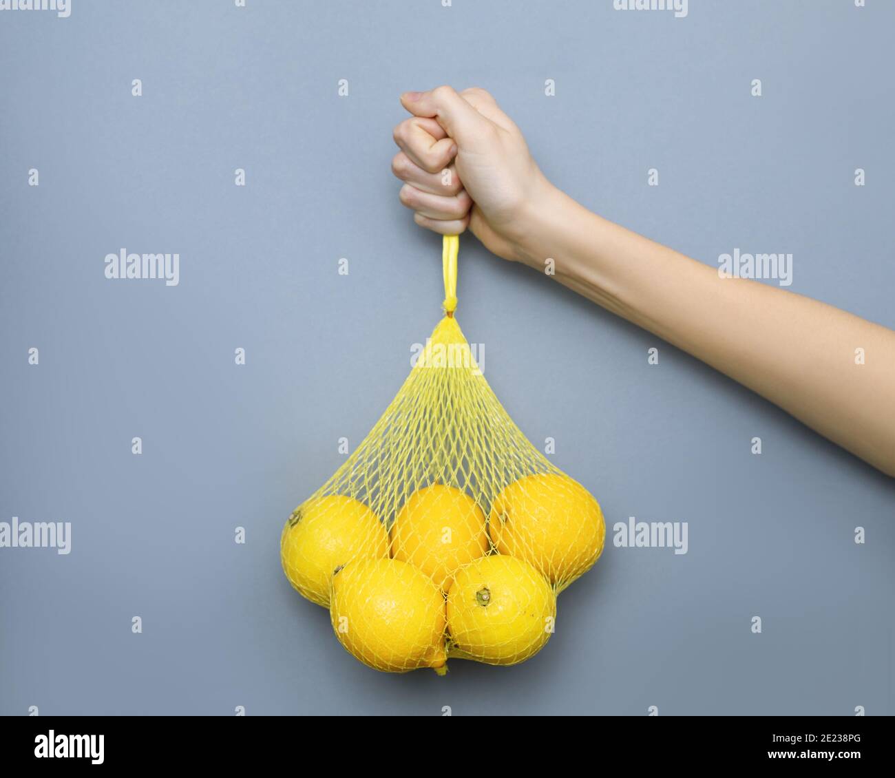 female hand holding yellow package mesh with lemons on gray background. Color concept 2021, healthy eating, moderate consumption Stock Photo