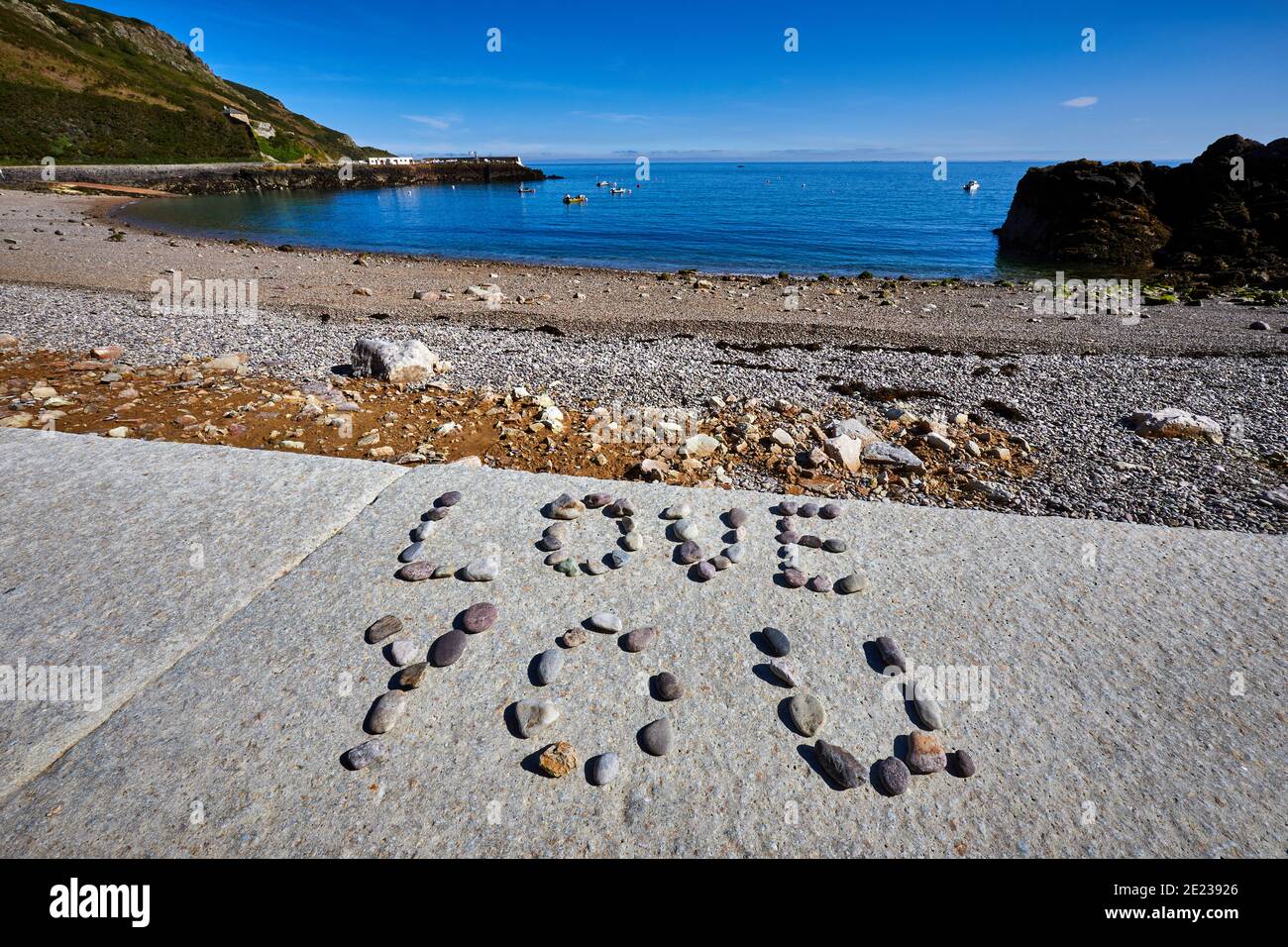 Image of a pebble beach in the early morning sunshine with a small island and harbour in the background with the words LOVE YOU written in pebbles in Stock Photo