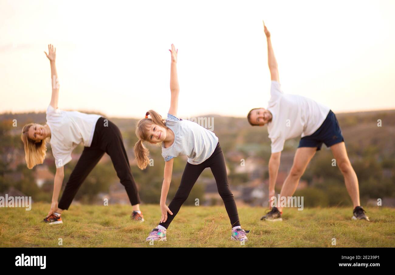 Parents instill sports habits in their child by example, focus on child.  Happy sporty family doing stretching exercises, practices yoga outdoor. Mom  dad and daughter doing sport exercises together Stock Photo -
