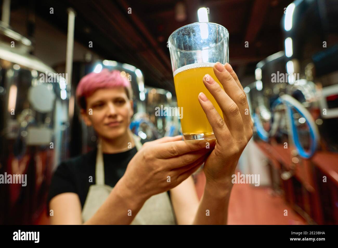 Hand of young female brewer in apron examining glass of fresh beer after preparation while standing among huge tanks in front of camera Stock Photo