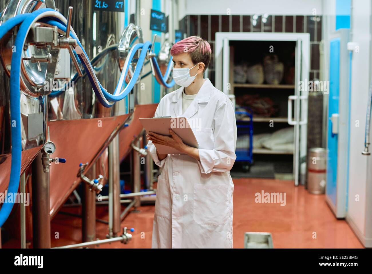 Young female brewery expert in whitecoat and protective mask making notes while looking at new steel equipment for beer production Stock Photo