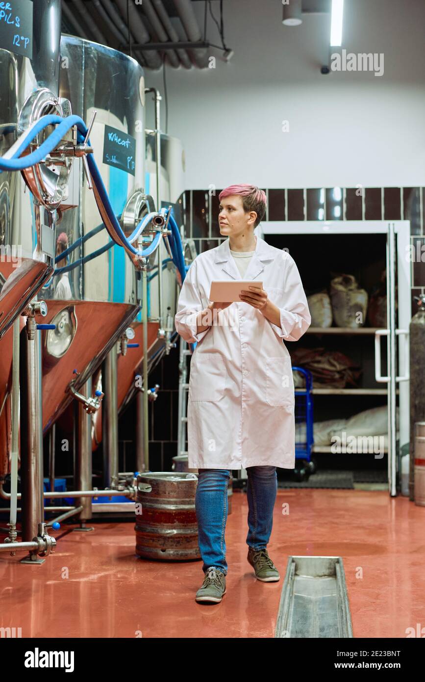 Young female brewery expert in whitecoat entering data in tablet while