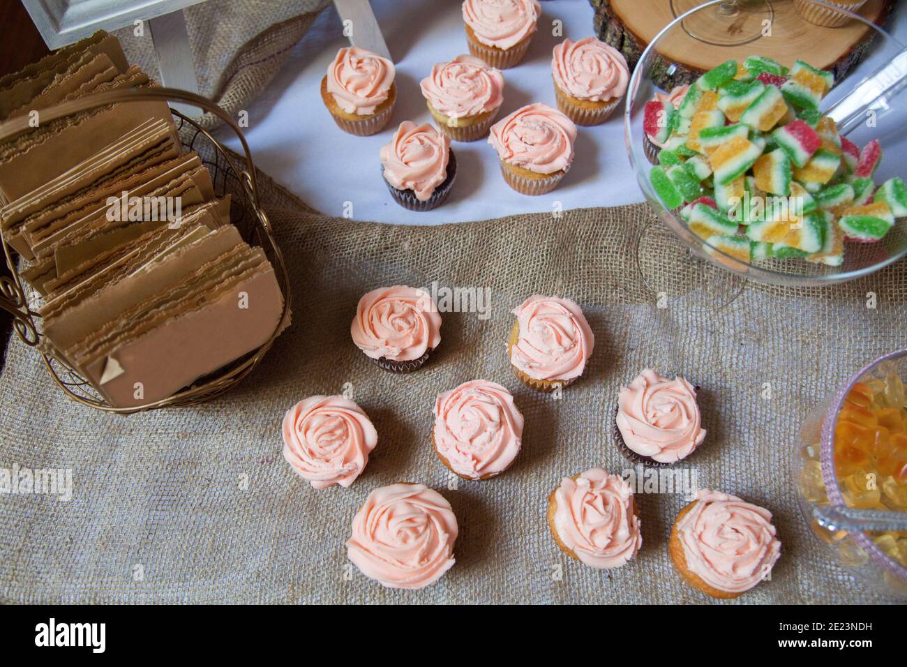 Pink frosted vanilla and chocolate cupcakes sit on a table cloth of burlap with other candies in the background as a dessert table for a wedding. Stock Photo