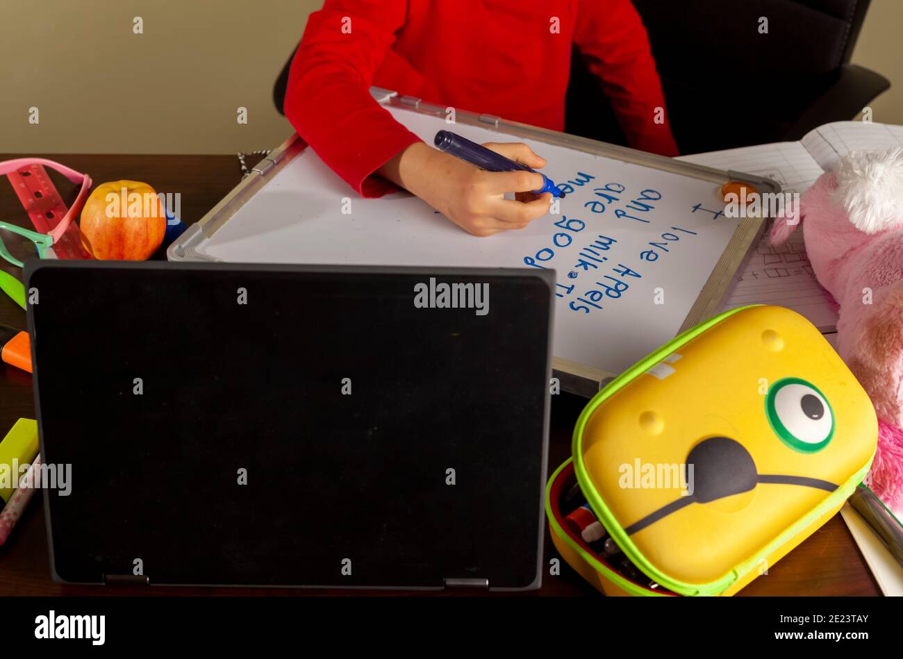 A cute little girl is writing sentences on a white board using blue marker. She is attending virtual classroom with her teacher through her laptop due Stock Photo