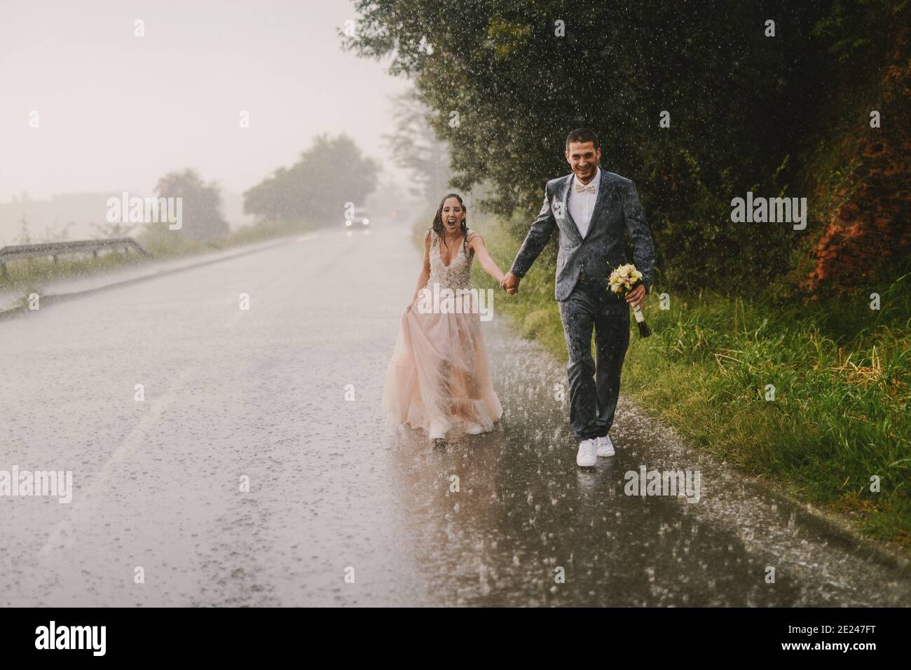 Just married couple holding hands and walking on rain. Walking in wet ceremonial clothes on driveway. Smiling and looking at camera. Stock Photo