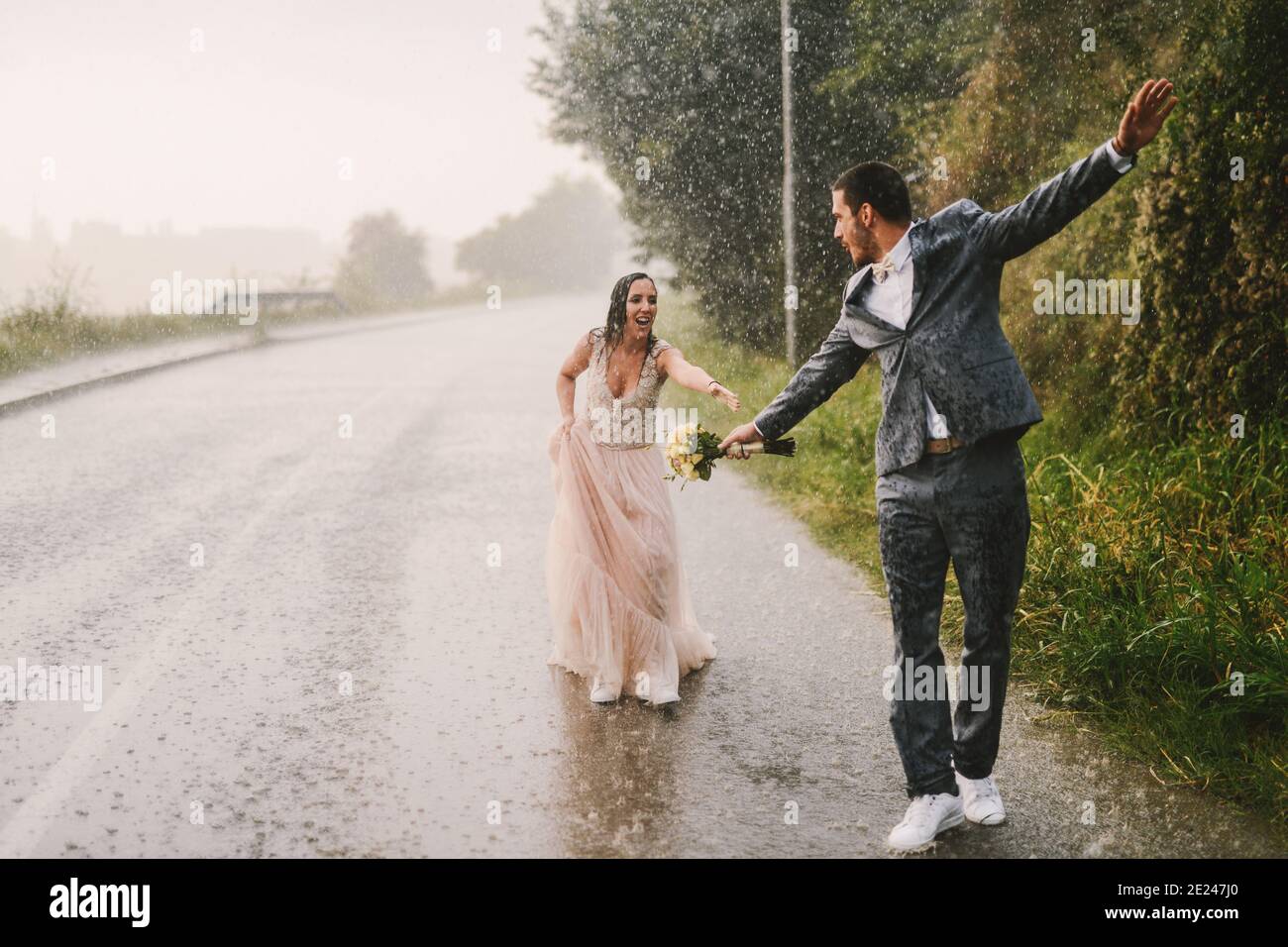 Totally wet just married couple walking on rain by drive road. Bride is train to catch her husband. Stock Photo