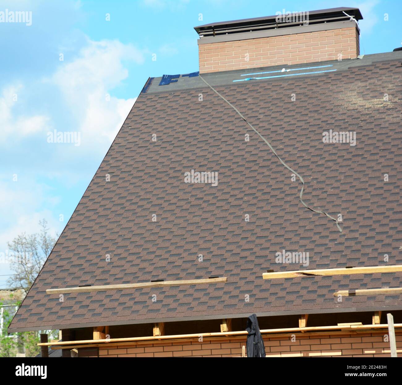 Incomplete roof with asphalt shingles installation on underlayment. Asphalt shingle tiles at the ridge of the rooftop near a chimney with flashing. Stock Photo