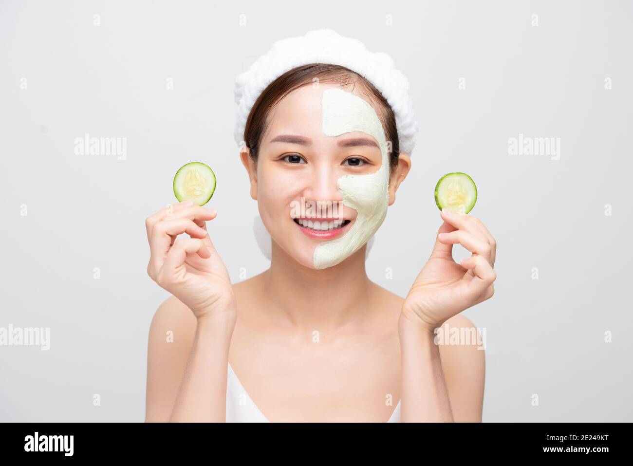 Beautiful young Asian woman with clay mask face holding cucumber piece over white background. Stock Photo