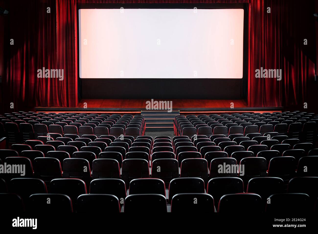 Auditorium of an empty movie theatre and stage with opened red velvet curtains viewed from the rear of the rows seats with lights on the screen Stock Photo