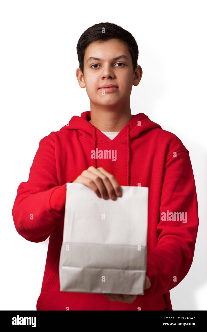 Teenage boy holding white paper bag in his hand isolated on white background. Young caucasian guy wearing red hoodie offering package with content Stock Photo