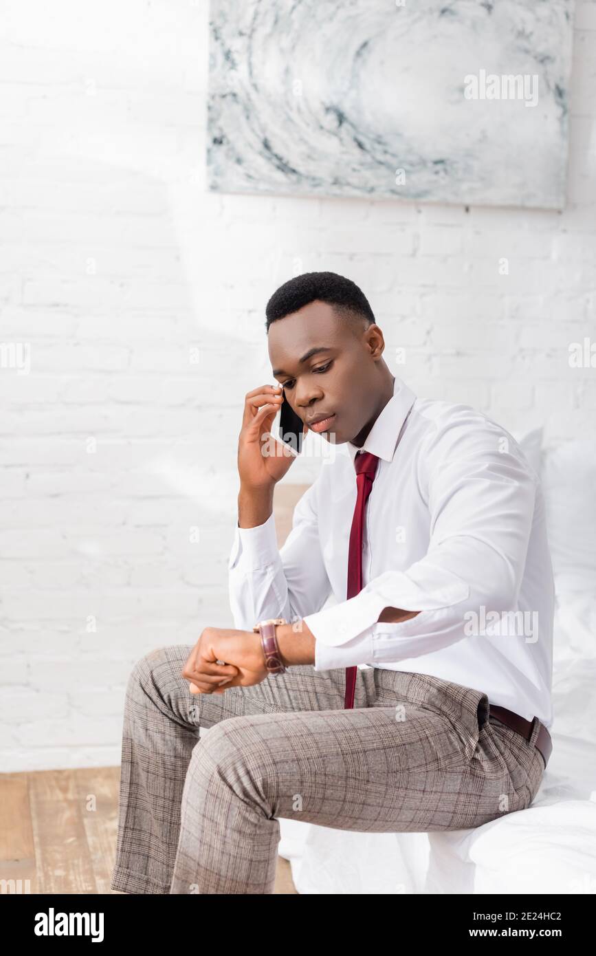African american businessman checking time while talking on smartphone on bed Stock Photo