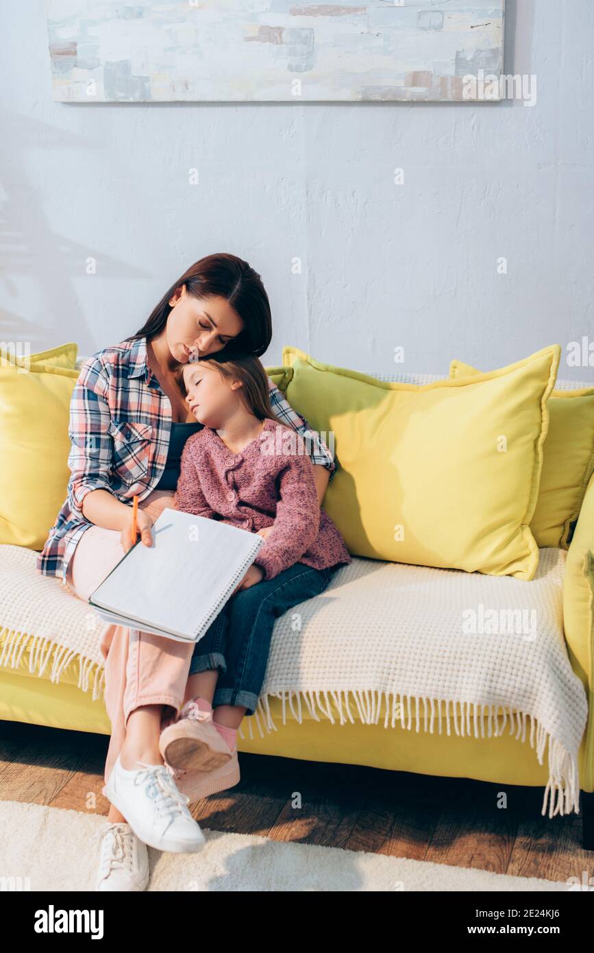 Full length of sad mother with copy book hugging daughter sleeping on couch at home Stock Photo